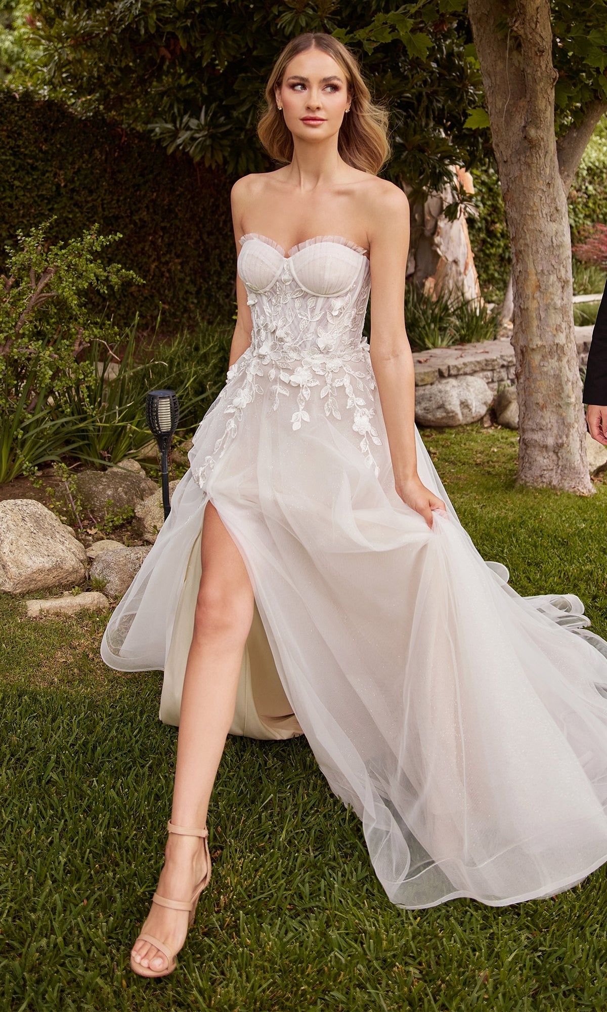 A woman in the Ladivine White Wedding Dress CD859W—an off-white, strapless gown with lace embroidery and a thigh-high slit—walks on grass outdoors.